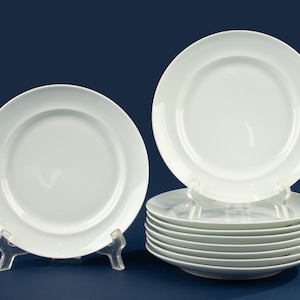 Puede incluir: Un juego de platos de cerámica blanca, exhibidos sobre soportes de plástico transparente sobre un fondo azul oscuro. Los platos tienen un diseño sencillo y elegante, con un borde ligeramente elevado. La imagen muestra una pila de platos y dos platos individuales.