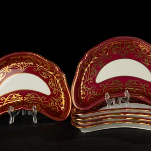 Puede incluir: Juego de cinco platos de servir de porcelana roja y dorada. Los platos tienen forma de media luna y tienen un centro blanco con un borde dorado.