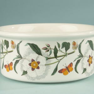 Op de afbeelding: Een ronde witte keramische kom met een herhalend botanisch patroon van grote witte bloemen met donkere harten, groene bladeren en oranje en gele vlinders die de buitenkant van de kom met een gladde rand rondom sieren.