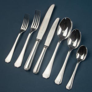 May include: A set of six polished silver-toned stainless steel flatware pieces arranged diagonally. The collection includes two forks, two knives with serrated blades, and three spoons of varying sizes, all featuring a classic handle design.
