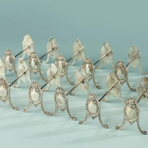 Op de afbeelding: Een verzameling zilverkleurige metalen messenleggers, elk voorzien van een sierlijke, ovale voet met gedetailleerde reliëfversieringen en een slanke, horizontale stang die vanuit het midden uitsteekt om bestek te ondersteunen.