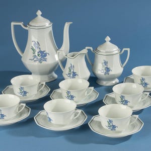 Może przedstawiać: Biały porcelanowy zestaw do herbaty składający się z wysokiego czajnika, dzbanuszka na mleko, cukiernicy z pokrywką oraz sześciu filiżanek z pasującymi spodkami. Każdy element ozdobiony jest niebiesko-zielonym motywem kwiatowym i cienką szarą obwódką.