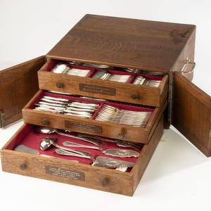 Può includere: Un portaposate in legno con tre cassetti, ciascuno foderato in velluto rosso e pieno di utensili d'argento. Il cofanetto è aperto, rivelando le forchette, i coltelli e i cucchiai ben disposti. L'esterno in legno ha una finitura naturale.
