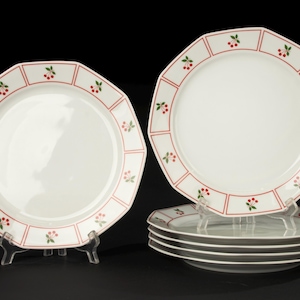 May include: A collection of white, octagonal plates with a red border, adorned with a repeating pattern of red cherries and green leaves. The plates are presented on clear stands against a black backdrop, showcasing a classic dinnerware design.