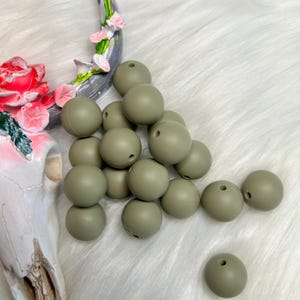 May include: A collection of olive green, round silicone beads with a small hole in each. The beads are scattered on a white, fluffy surface. A decorative skull and floral accent are visible in the background.