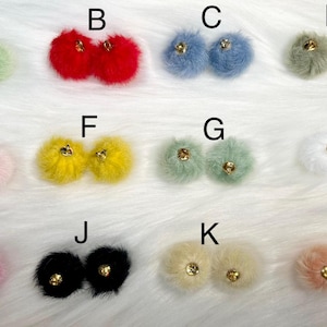 May include: A collection of fluffy pom-pom earrings in various colors, including green, red, blue, gray, pink, yellow, white, black, and peach. Each pair features a gold-colored stud. The earrings are arranged on a white, textured surface.