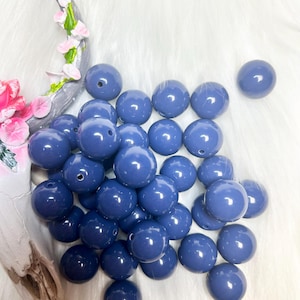 May include: A collection of glossy, periwinkle blue beads. The round beads have a hole drilled through the center. The beads are scattered on a white, fluffy surface. A decorative piece with pink and green floral accents is in the background.