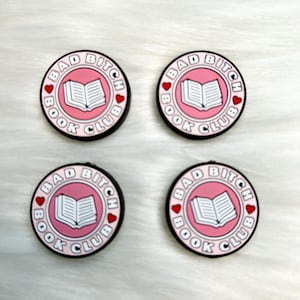 May include: Four round, black and pink accessories with the text "BAD BITCH BOOK CLUB" encircling a white book graphic. Each accessory has a pink center, a black border, and small red heart accents.