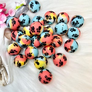 May include: A collection of colorful round beads with a paw print design. The beads are in shades of blue, pink, yellow, and orange. The paw prints are black. The beads are scattered on a white, fluffy surface.