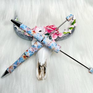 May include: A white pen with a floral pattern and a silver tip. The pen is lying on a white skull with floral accents.