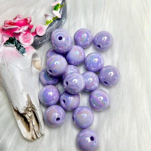 May include: A pile of iridescent lavender beads with a white, iridescent finish. The beads are round and have a hole in the center.