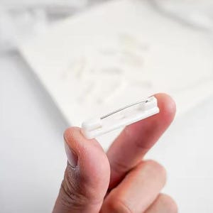 May include: A white plastic safety pin with a silver metal pin. The pin is held in a person's hand.