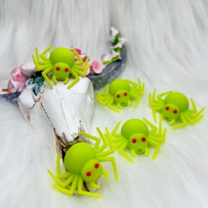May include: A collection of lime green spider figurines with red eyes and eight legs. The spiders are arranged on a white surface, with a decorative skull and floral arrangement in the background. These Halloween-themed decorations are perfect for adding a spooky touch.