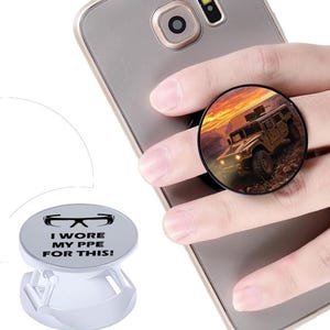 Puede incluir: Un soporte circular para teléfono con un gráfico de un vehículo militar color canela al atardecer. Un segundo soporte blanco se muestra por separado, con gafas de seguridad negras y el texto I WORE MY PPE FOR THIS! en letras negras.