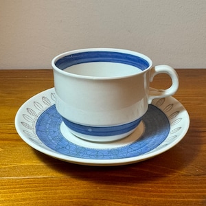 Może przedstawiać: Biały kubek do herbaty i spodek z niebiesko-białym geometrycznym wzorem. Kubek ma niebieską obwódkę wokół górnej i dolnej części.