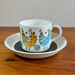 May include: A white ceramic coffee cup and saucer with a black and white bird design. The birds are stylized and have yellow, blue, and black accents.