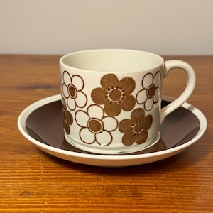 May include: A white ceramic mug with a black handle and the text "But First, Coffee" in a bold, black font. The mug is filled with a dark liquid, likely coffee, and sits on a light-colored surface.