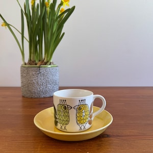 May include: A white ceramic coffee cup with a yellow saucer. The cup features a whimsical design of two stylized birds in yellow, green, and black. A potted plant with yellow daffodils is in the background.