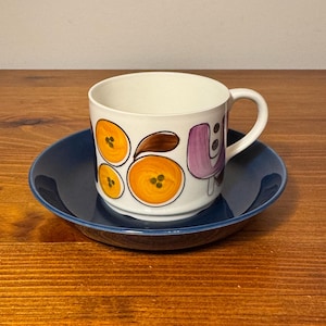 May include: A white ceramic mug with a black handle and the text "But First, Coffee" in a black, bold font. The mug is sitting on a wooden surface. The mug is approximately 4 inches tall.