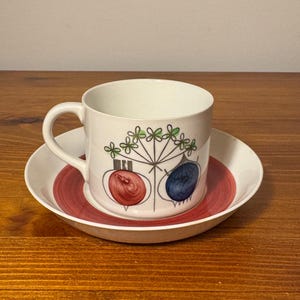 May include: A white ceramic teacup and saucer set. The cup and saucer have a red and white color scheme. The cup features a design with red and blue bulb-shaped figures and green floral accents. The set is placed on a wooden surface.