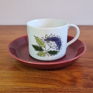 May include: A white ceramic teacup with a floral design in shades of green, purple, and white. The cup has a handle and is sitting on a burgundy saucer.