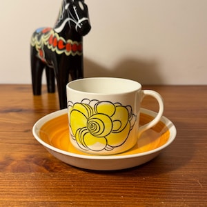 May include: A white ceramic mug with a black handle and the text "But First, Coffee" in a bold, black font. The mug is set against a blurred background, suggesting a cozy setting.