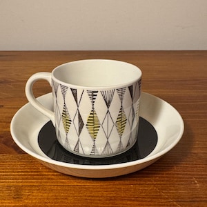 May include: A white teacup with a handle and matching saucer. The cup and saucer have a black, yellow, and white geometric diamond pattern. The cup sits on a black circle in the center of the saucer. The set is on a wooden surface.