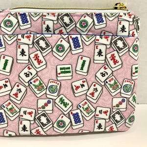 May include: A pink zippered pouch with a white background and a pattern of mahjong tiles. The tiles have Chinese characters and numbers on them.