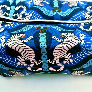 May include: A rectangular fabric pouch featuring a repeating pattern of white tigers with black stripes and red accents, set against a dark blue background with light blue tropical palm leaves and foliage, finished with a silver-toned metal zipper.