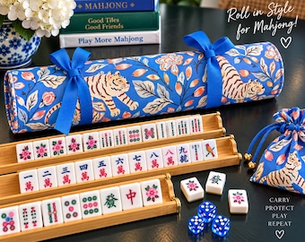 May include: A blue fabric roll with a tiger and floral pattern tied with blue ribbons, three bamboo tile racks holding white mahjong tiles with colorful symbols, a matching drawstring pouch, and three blue translucent dice on a surface.