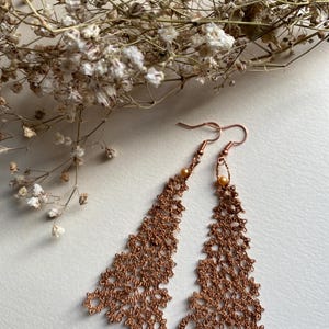 Può includere: Un paio di orecchini color rame con un delicato e intricato design in pizzo. Gli orecchini sono di forma triangolare e presentano una piccola perla rotonda nella parte superiore.
