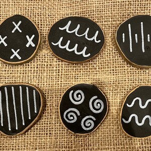 Puede incluir: Una colecci&oacute;n de diez posavasos redondos de madera negra con dise&ntilde;os pintados en blanco. Cada posavasos presenta un patr&oacute;n &uacute;nico, que incluye flechas, cruces, l&iacute;neas onduladas y espirales. Los posavasos se exhiben sobre una superficie de tela texturizada de color beige.
