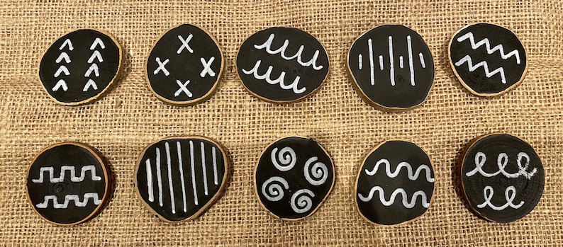 Puede incluir: Una colecci&oacute;n de diez posavasos redondos de madera negra con dise&ntilde;os pintados en blanco. Cada posavasos presenta un patr&oacute;n &uacute;nico, que incluye flechas, cruces, l&iacute;neas onduladas y espirales. Los posavasos se exhiben sobre una superficie de tela texturizada de color beige.