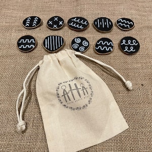 Puede incluir: Un conjunto de diez discos de madera negros con dise&ntilde;os geom&eacute;tricos blancos, incluyendo l&iacute;neas y ondas. Una bolsa con cord&oacute;n con el texto "Early Years Play" se encuentra en primer plano, sobre una superficie de arpillera.