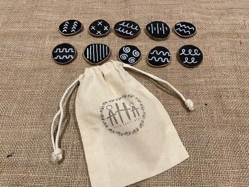 Puede incluir: Un conjunto de diez discos de madera negros con dise&ntilde;os geom&eacute;tricos blancos, incluyendo l&iacute;neas y ondas. Una bolsa con cord&oacute;n con el texto "Early Years Play" se encuentra en primer plano, sobre una superficie de arpillera.