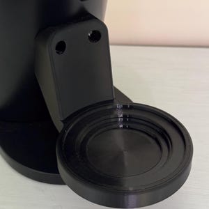 May include: A black coffee maker with a circular foot pedal. The foot pedal has a textured surface with concentric circles.