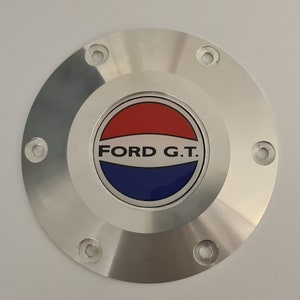 Op de afbeelding: Een rond zilveren auto-embleem met een rood, wit en blauw logo met de tekst "FORD G.T."