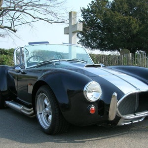 May include: A black and silver classic sports car with a white stripe down the bonnet. The car is parked in front of a stone cross and a tree.