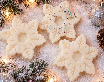 Puede incluir: Tres fundentes de cera de color blanco roto con forma de copo de nieve y patrones geométricos en relieve. Una pieza presenta destellos iridiscentes. Las figuras están dispuestas sobre nieve artificial, rodeadas de ramas de pino y bayas rojas.
