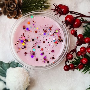 May include: A pink wax melt with a glittery finish in a clear container. The wax melt is surrounded by a white, fluffy surface and green pine branches with red berries.