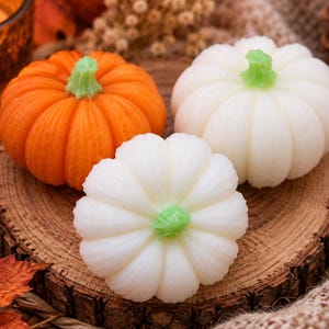 May include: Three small pumpkin-shaped candles, one in vibrant orange and two in off-white, each featuring a distinct ribbed texture and a small, light green stem at the top. The candles are arranged together, showcasing their rounded, realistic forms.
