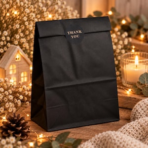 May include: A matte black paper gift bag with a folded top edge, sealed with a circular black sticker featuring the text THANK YOU in gold lettering. The bag has a rectangular base and stands upright with a clean, minimalist design.