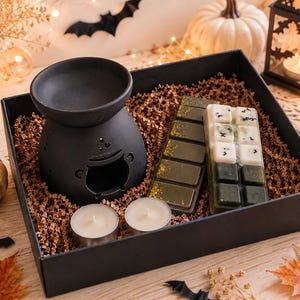 May include: A black ceramic cauldron-shaped wax warmer, two round tea light candles, and two rectangular wax melt bars with green and white segments are arranged inside a black gift box filled with brown paper crinkle shred filler.