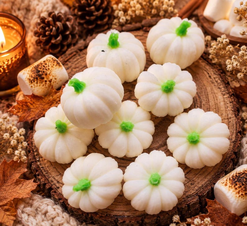 May include: A collection of small, white, pumpkin-shaped sweets with bright green stems. The sweets are arranged on a circular wooden slice, surrounded by toasted marshmallows, dried flowers, and pinecones, with a lit candle nearby.