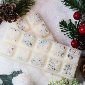 May include: A white wax melt with silver glitter, shaped like a bar of chocolate, with 12 individual squares. The wax melt is on a white surface with green pine branches and a pine cone.