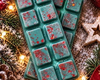 Puede incluir: Dos barras rectangulares de cera aromática en un color verde azulado profundo, cada una dividida en diez segmentos cuadrados. La superficie de la cera tiene una textura con purpurina roja dispersa y pequeñas motas rojas, creando un aspecto moteado.