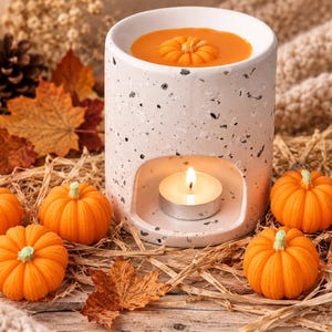 May include: A cylindrical white ceramic wax warmer with a speckled terrazzo pattern, featuring a hollow base for a tea light candle and a top basin filled with melted orange wax shaped like a small pumpkin, surrounded by miniature orange pumpkins.