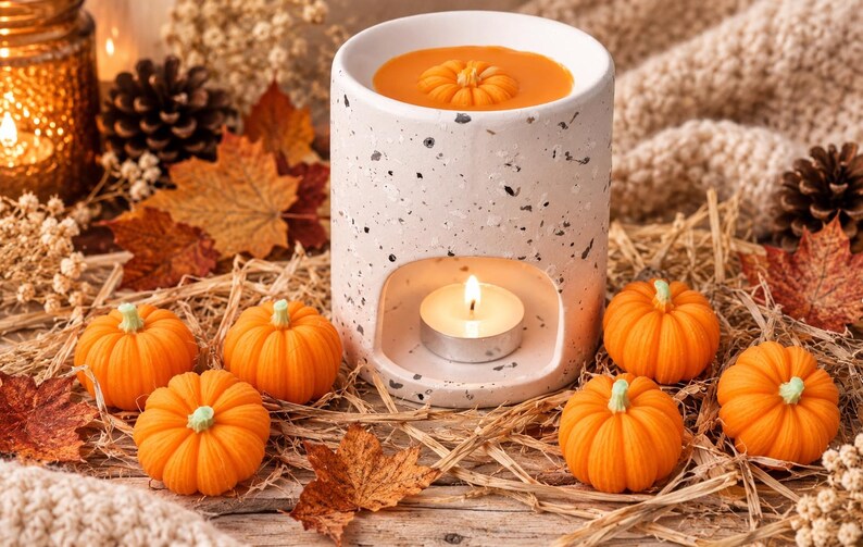 May include: A cylindrical white ceramic wax warmer with a speckled terrazzo pattern, featuring a hollow base for a tea light candle and a top basin filled with melted orange wax shaped like a small pumpkin, surrounded by miniature orange pumpkins.