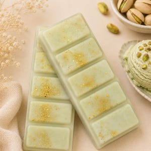 May include: Two rectangular wax melt bars in a pale sage green colour, each divided into five segments. The surface of the wax is decorated with scattered gold glitter particles that catch the light, creating a shimmering effect across the smooth finish.