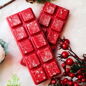 May include: Two red wax melts with a glittery finish, shaped like a bar of chocolate, with a festive holiday theme.